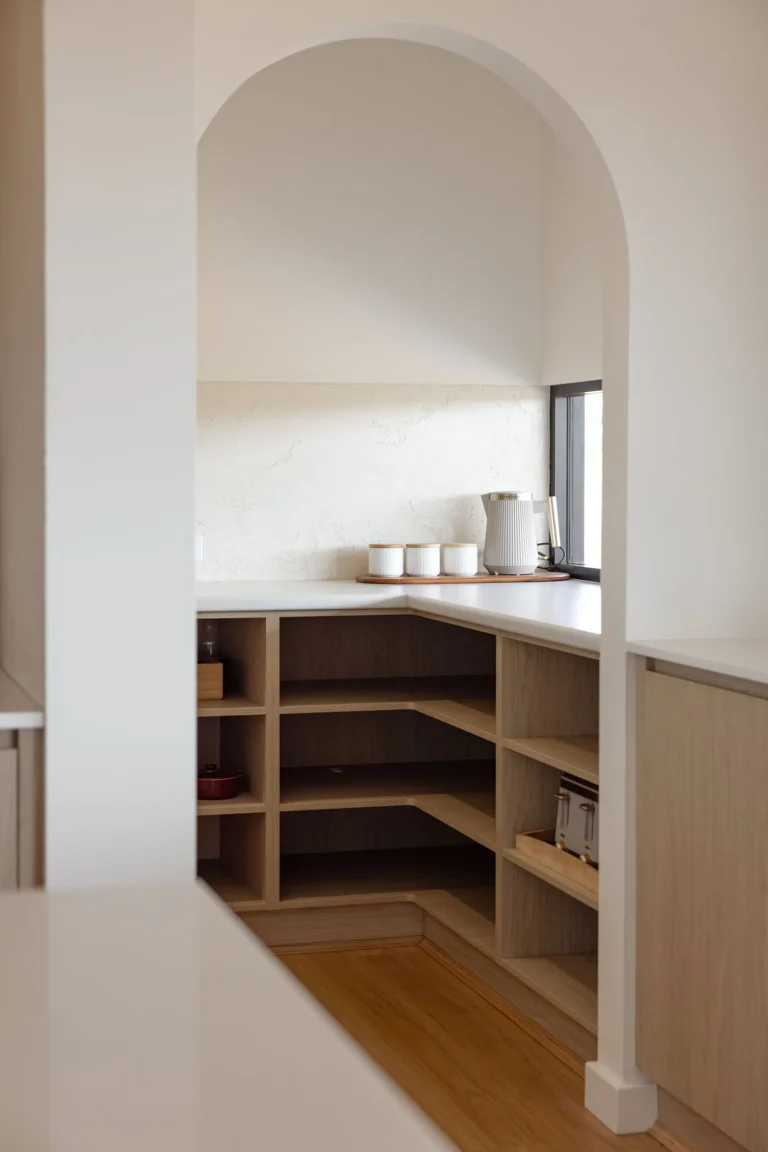 Arched opening into the butler's pantry with open oak-look shelving, a stone benchtop and textured render splashback