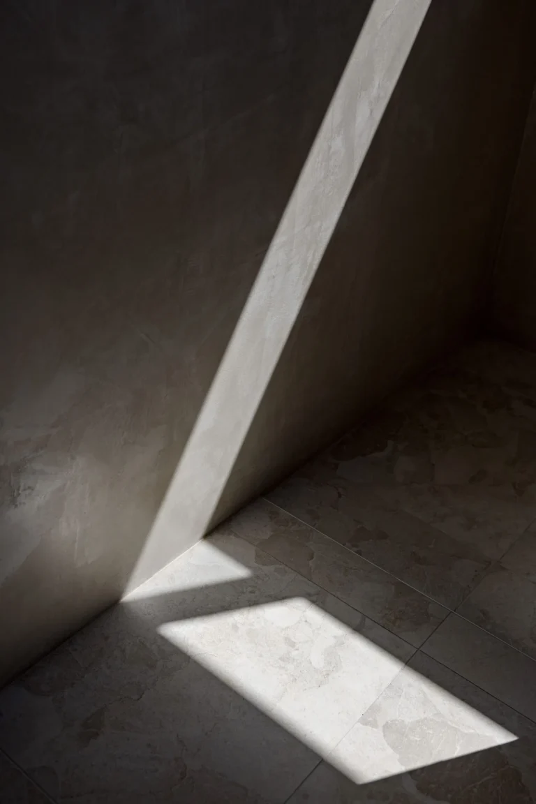 Shaft of sunlight across the rendered walls and stone-look floor tiles of the ensuite