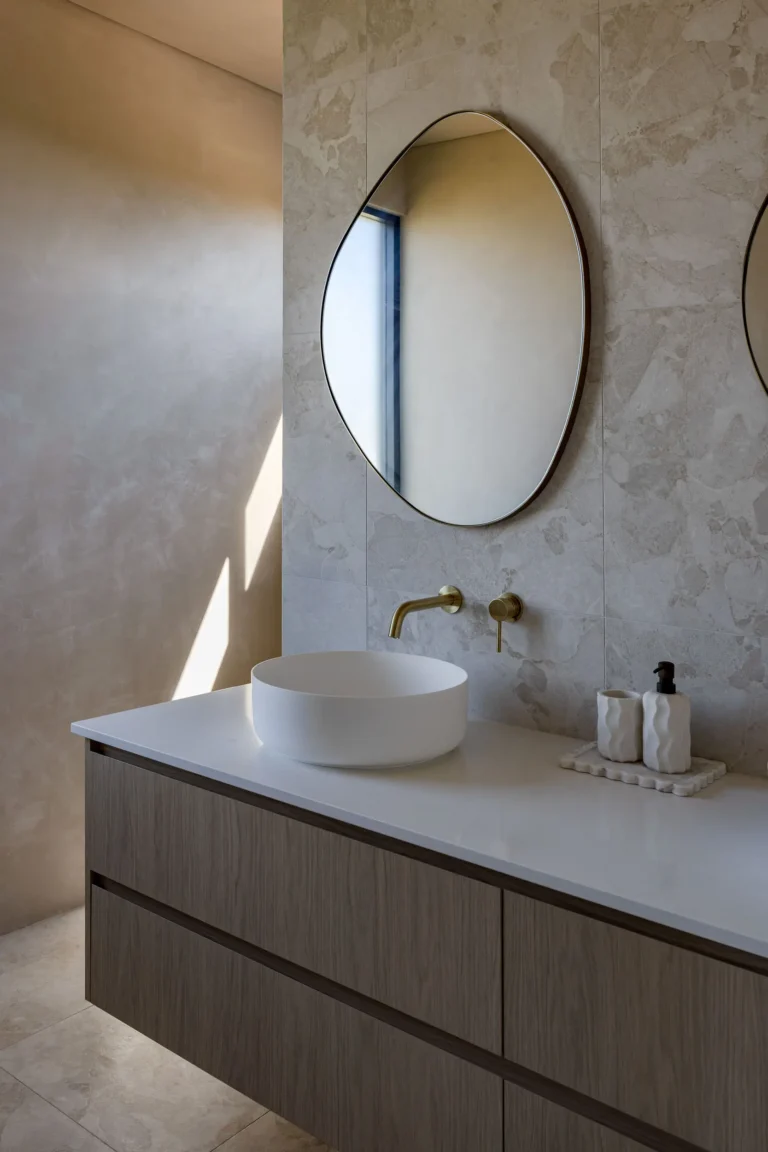 Ensuite vanity at Felling Karridale with a white vessel basin, brushed brass wall mixer, pebble-shaped mirror and timber-look floating cabinetry against stone-look tiles