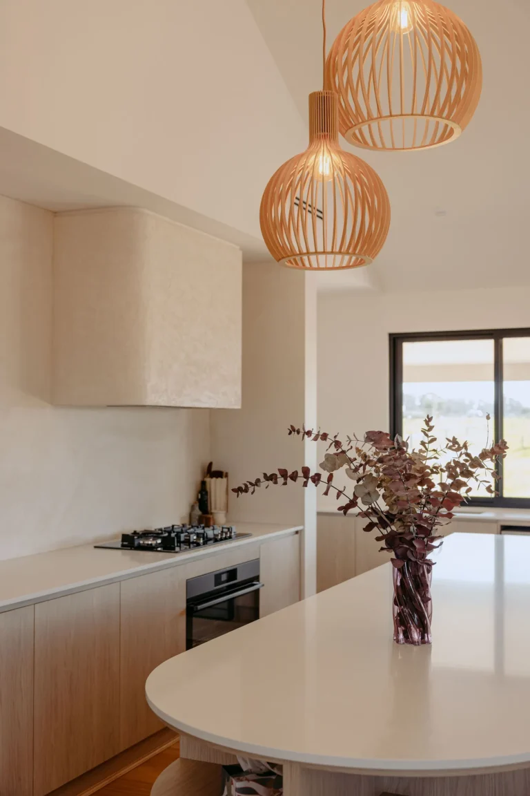 Curved stone island benchtop with a vase of dried eucalyptus under timber pendant lights, looking toward the kitchen window