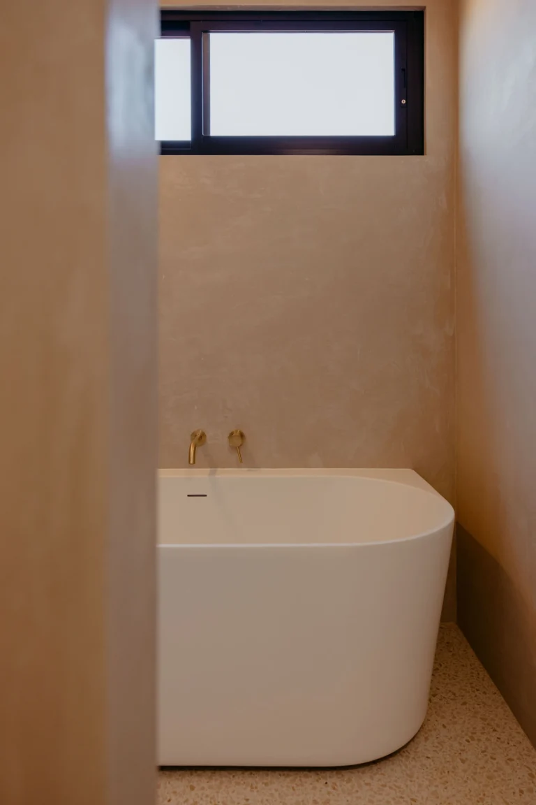 Back-to-wall freestanding bath with a brushed brass wall spout, rendered walls and terrazzo floor beneath a highlight window