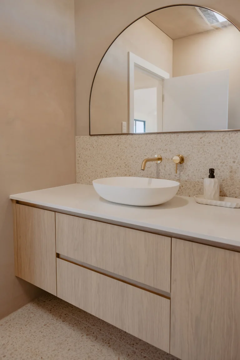 Floating oak-look vanity with an oval vessel basin, brushed brass wall mixer and terrazzo splashback beneath an arched mirror