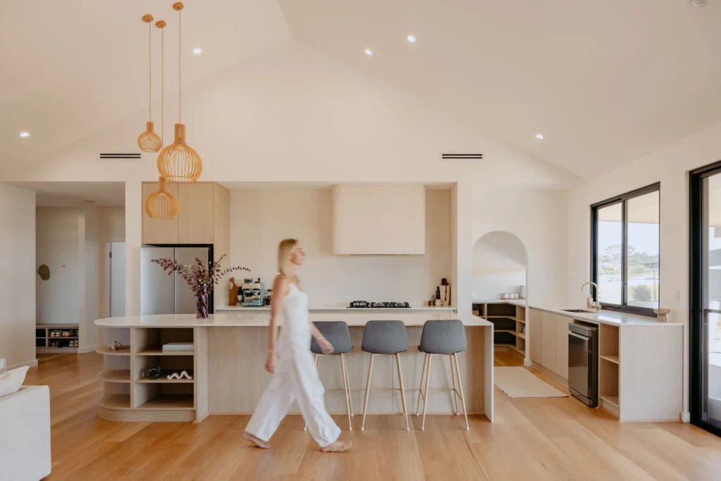 Open-plan kitchen at Felling Karridale with a raked ceiling, timber cage pendants over a curved-end island, light oak-look joinery and an arched butler's pantry beyond