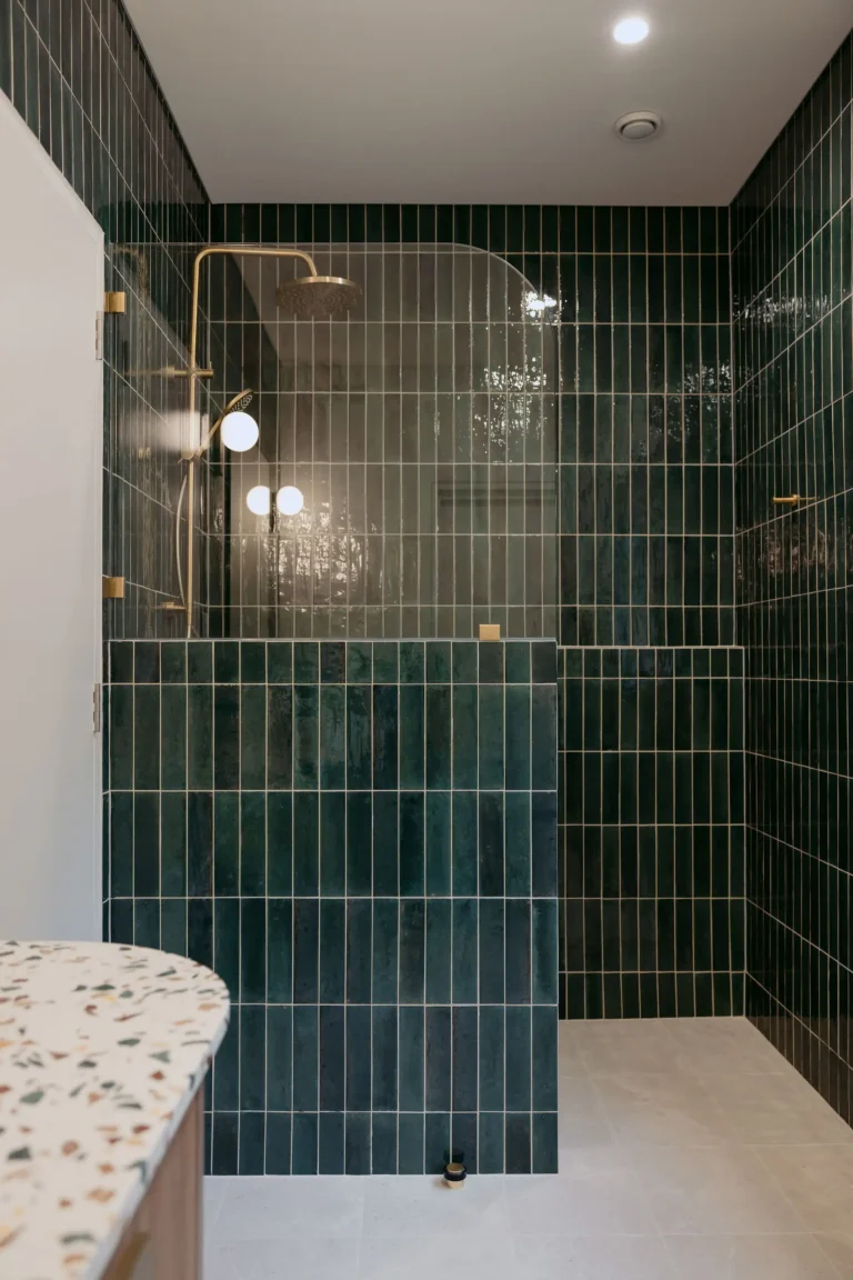 Walk-in shower with vertical dark green tiles, brass fixtures and a terrazzo vanity in the foreground