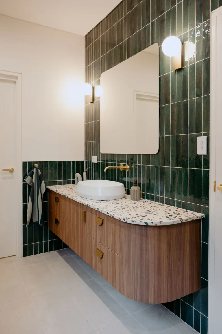 Bathroom with a green tiled feature wall, curved timber vanity, terrazzo top and round mirror with globe wall lights