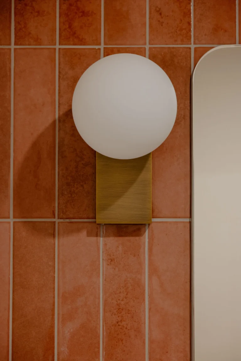 Frosted glass globe wall light on a brass mount against terracotta tiles beside a mirror