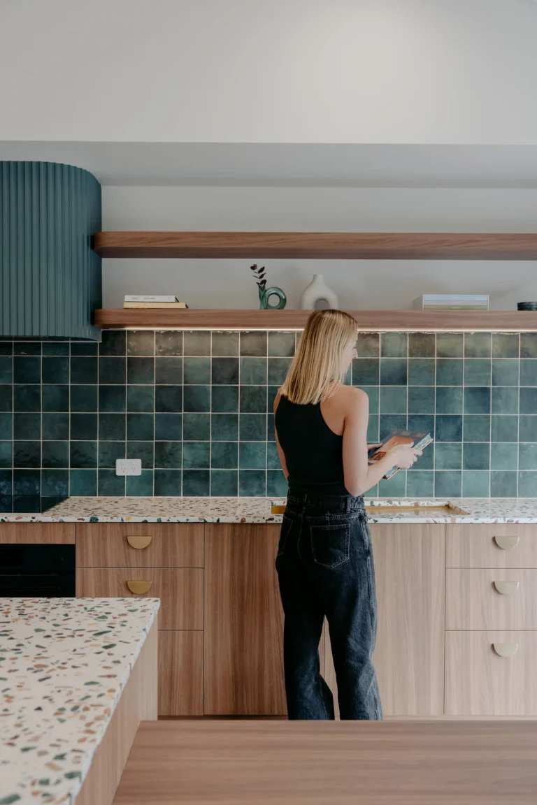 Woman standing at a modern kitchen bench with a green tiled splashback, terrazzo island and timber cabinetry with brass handles