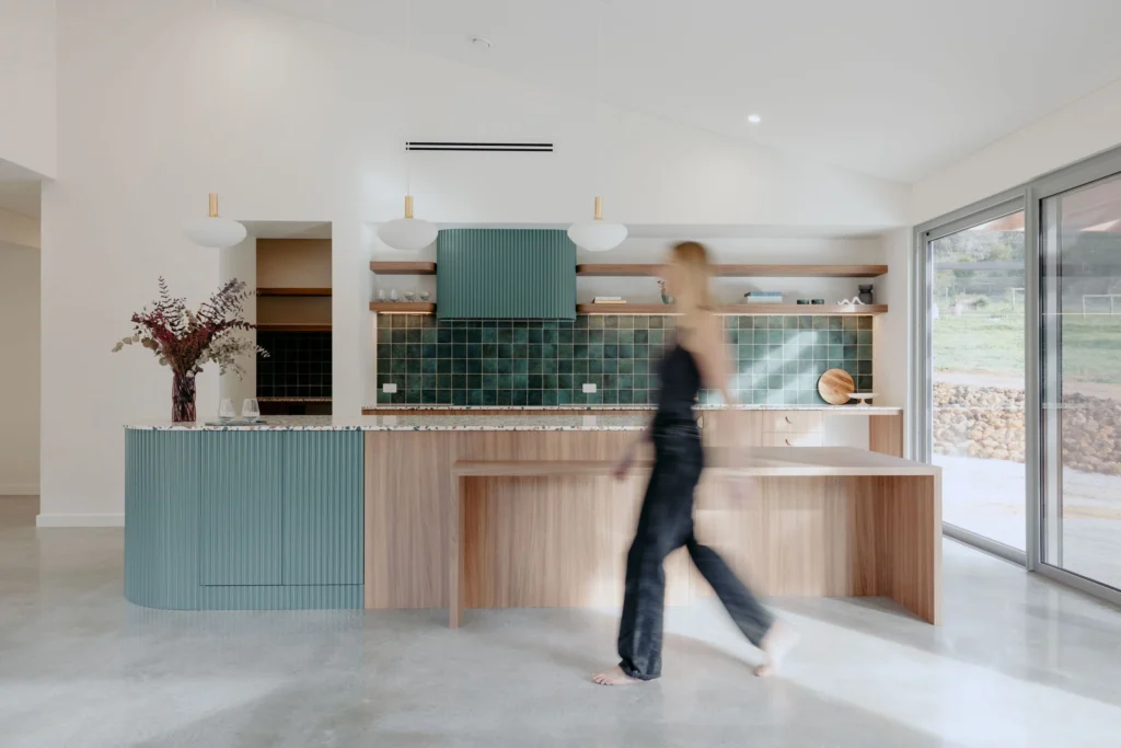 Open-plan kitchen and living area with a vaulted ceiling and sage-green fluted island bench opening onto bushland