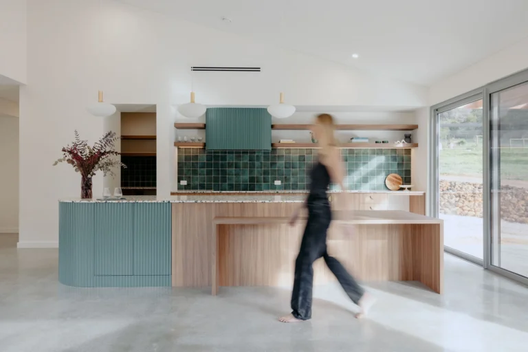 Open-plan kitchen and living area with a vaulted ceiling and sage-green fluted island bench opening onto bushland
