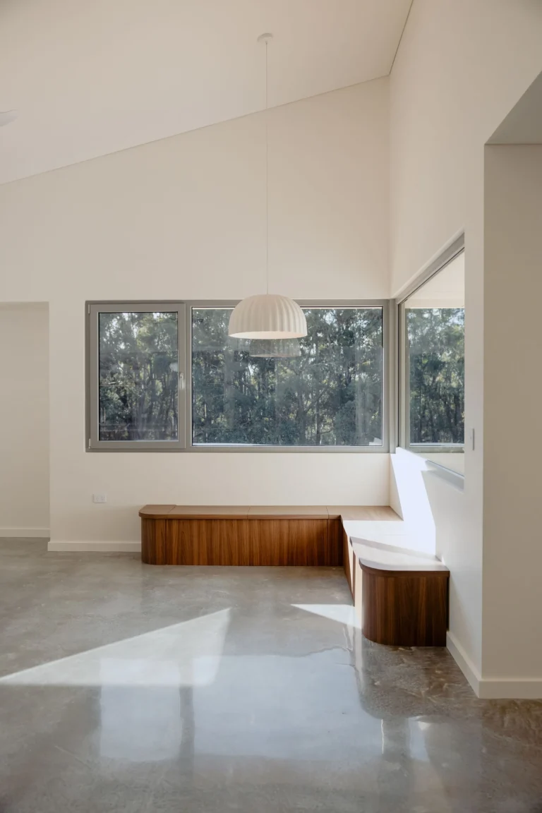 Living space with a raked ceiling, corner windows and a built-in timber bench seat overlooking native bush