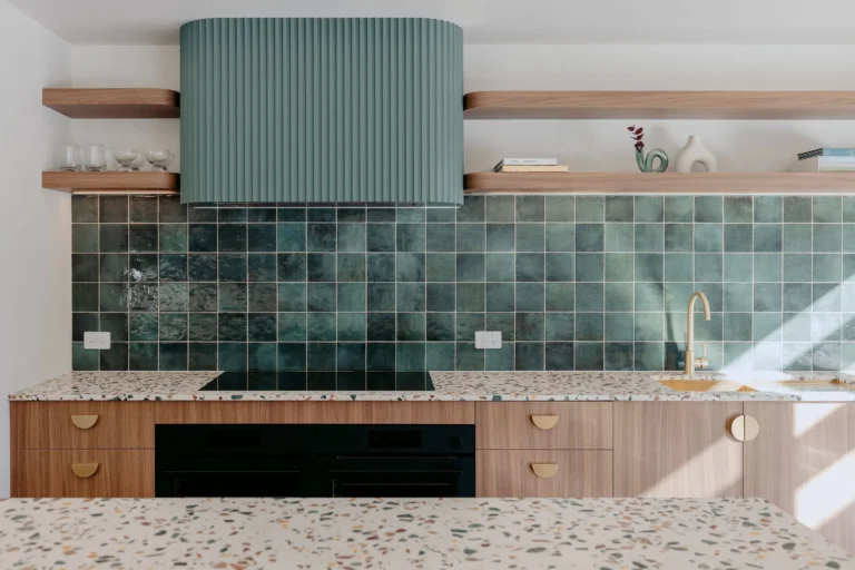 Kitchen detail with a green glazed tile splashback, fluted sage-green rangehood, timber shelves and terrazzo benchtop