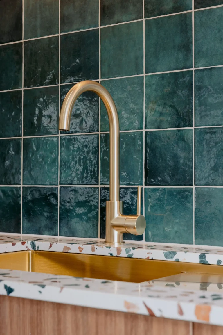 Brushed brass gooseneck mixer tap over a brass sink with a green square-tiled splashback