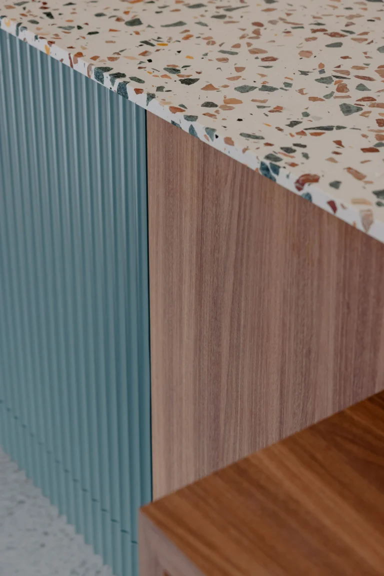 Detail of a terrazzo benchtop edge meeting teal fluted joinery and timber cabinetry