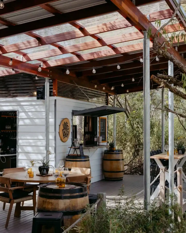 Valle du Venom cellar door deck in Yallingup showing round timber dining table, wine barrel furniture, tasting bar, and branded logo signage surrounded by Australian bushland
