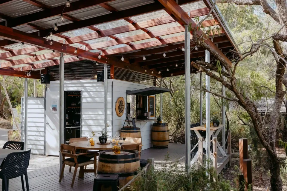 Valle du Venom cellar door deck in Yallingup showing round timber dining table, wine barrel furniture, tasting bar, and branded logo signage surrounded by Australian bushland