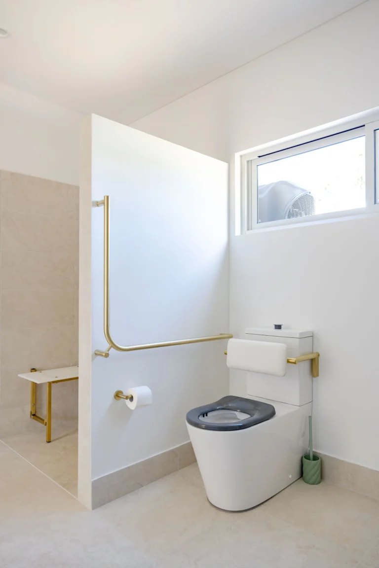 Accessible toilet with brushed brass grab rails and backrest beside a step-free shower with bench seat