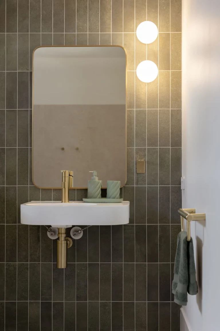 Softly lit bathroom vanity with globe wall lights, brass-framed mirror and sage green tiled wall