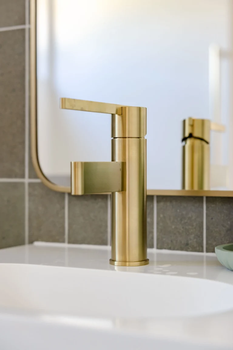 Detail of a brushed brass basin mixer on a white wall-hung basin with sage tiles behind